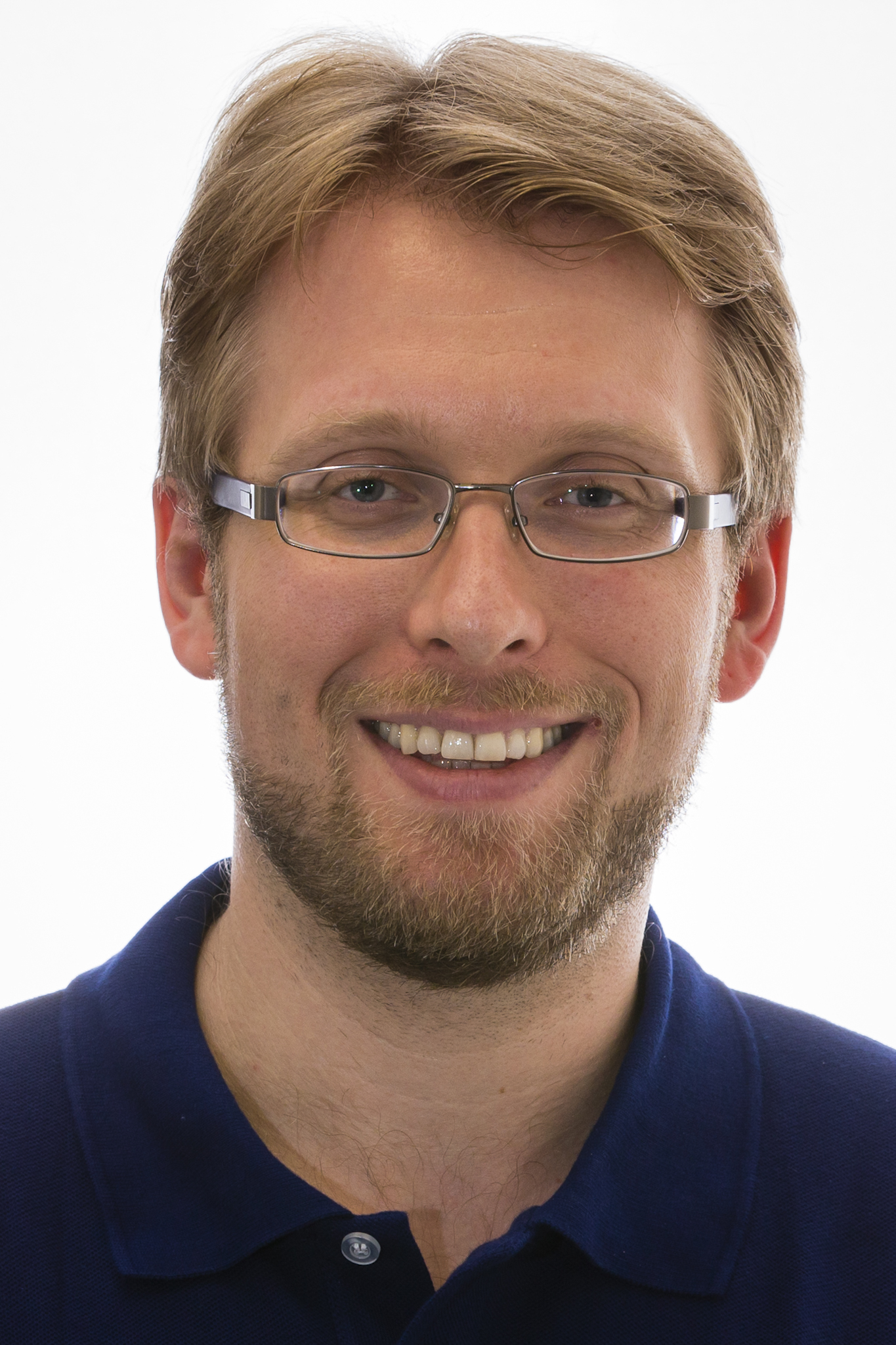 A headshot of smiling Jan Gruber who has short light brown hair, a light brown mustache, and a light brown beard. He is wearing glasses with a grey frame and a dark blue polo shirt.