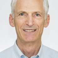 A headshot of smiling A J (Jon) Berrick who has short white hair, wearing a light blue shirt.