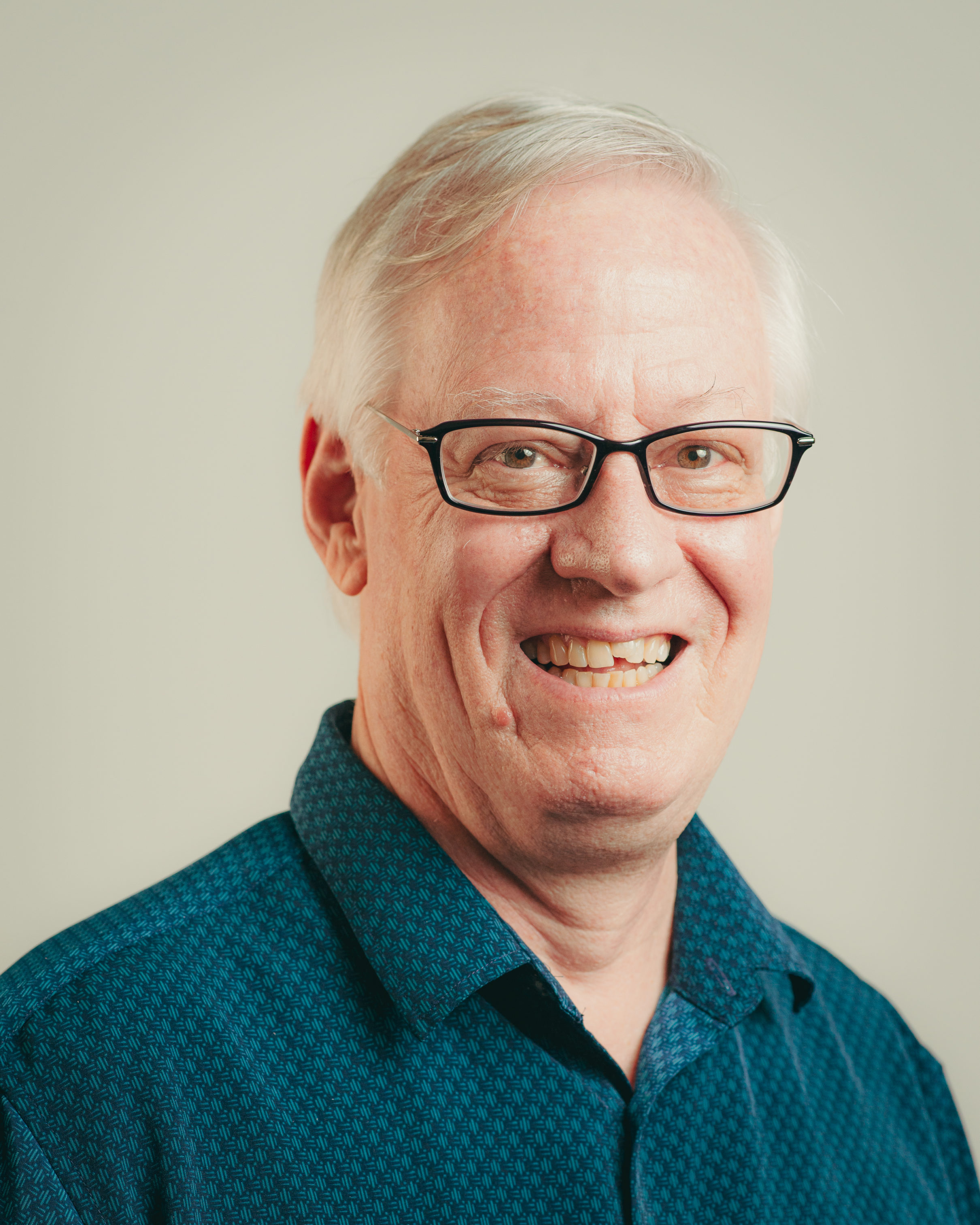 A headshot of smiling Michael F Maniates who has short white hair, is wearing glasses with a black frame, and a bright teal collared shirt.