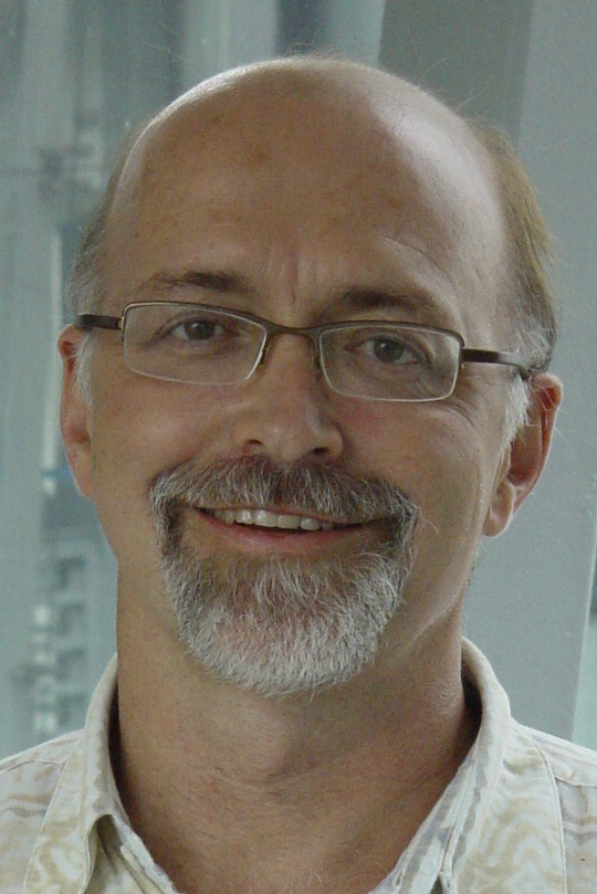 A headshot of smiling Neil Clarke who has salt and pepper hair and is wearing glasses with a brown frame. He is also wearing a cream collared shirt and is standing in front of a glass window.