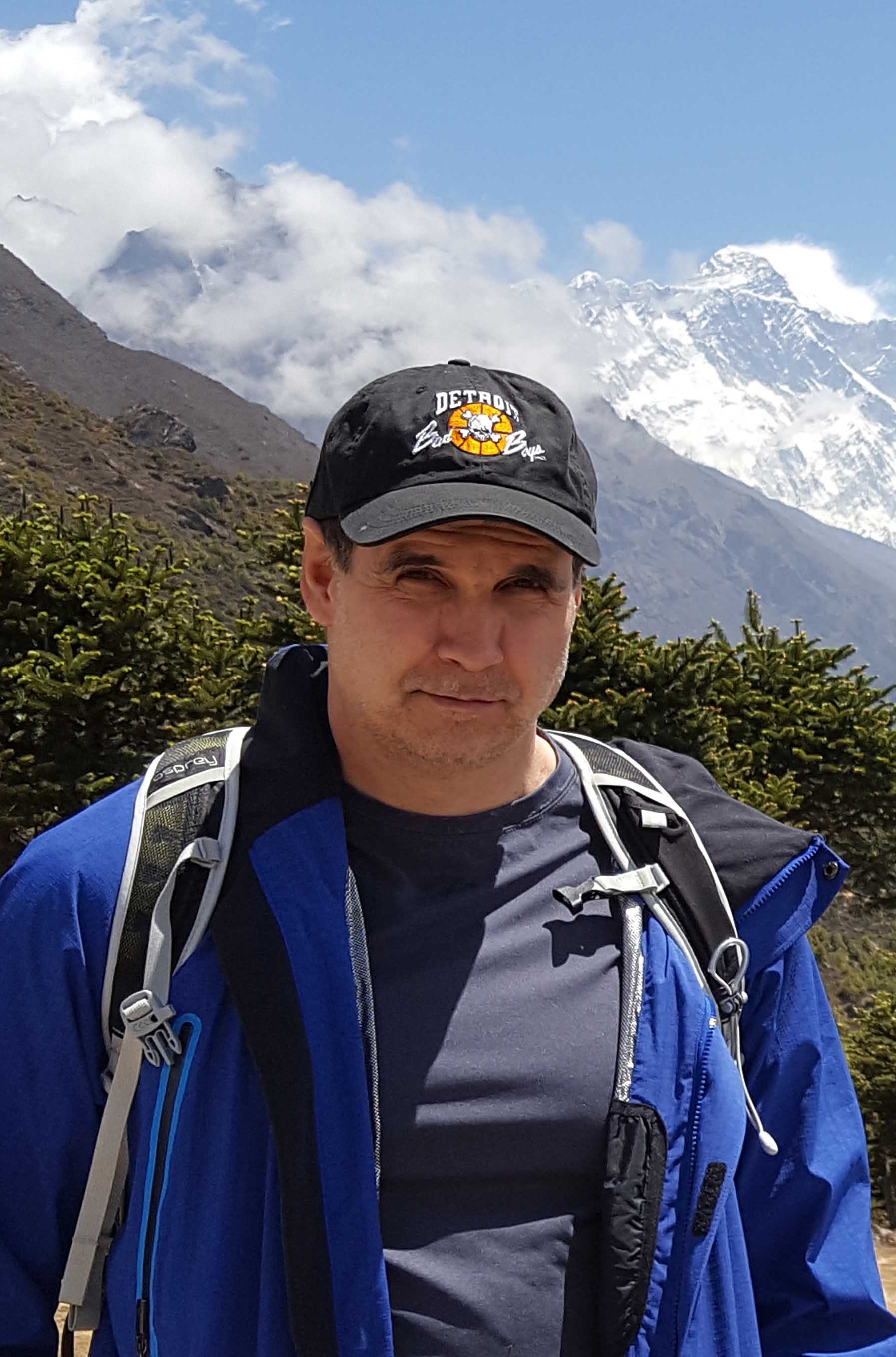 A headshot of smiling Scott Cook who has short brown hair, wearing a black baseball cap that reads “Detroit Bad Boys”. He is also wearing a blue t-shirt and a blue windbreaker and a black backpack. He is posing in front of snow-capped mountains and greenery.