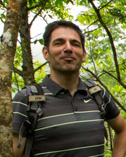 A headshot of smiling Michiel van Breugel who has short black hair. He is wearing a brown Nike shirt with horizontal stripes in grey and white. He is carrying a backpack and is standing in front of trees in a forest.