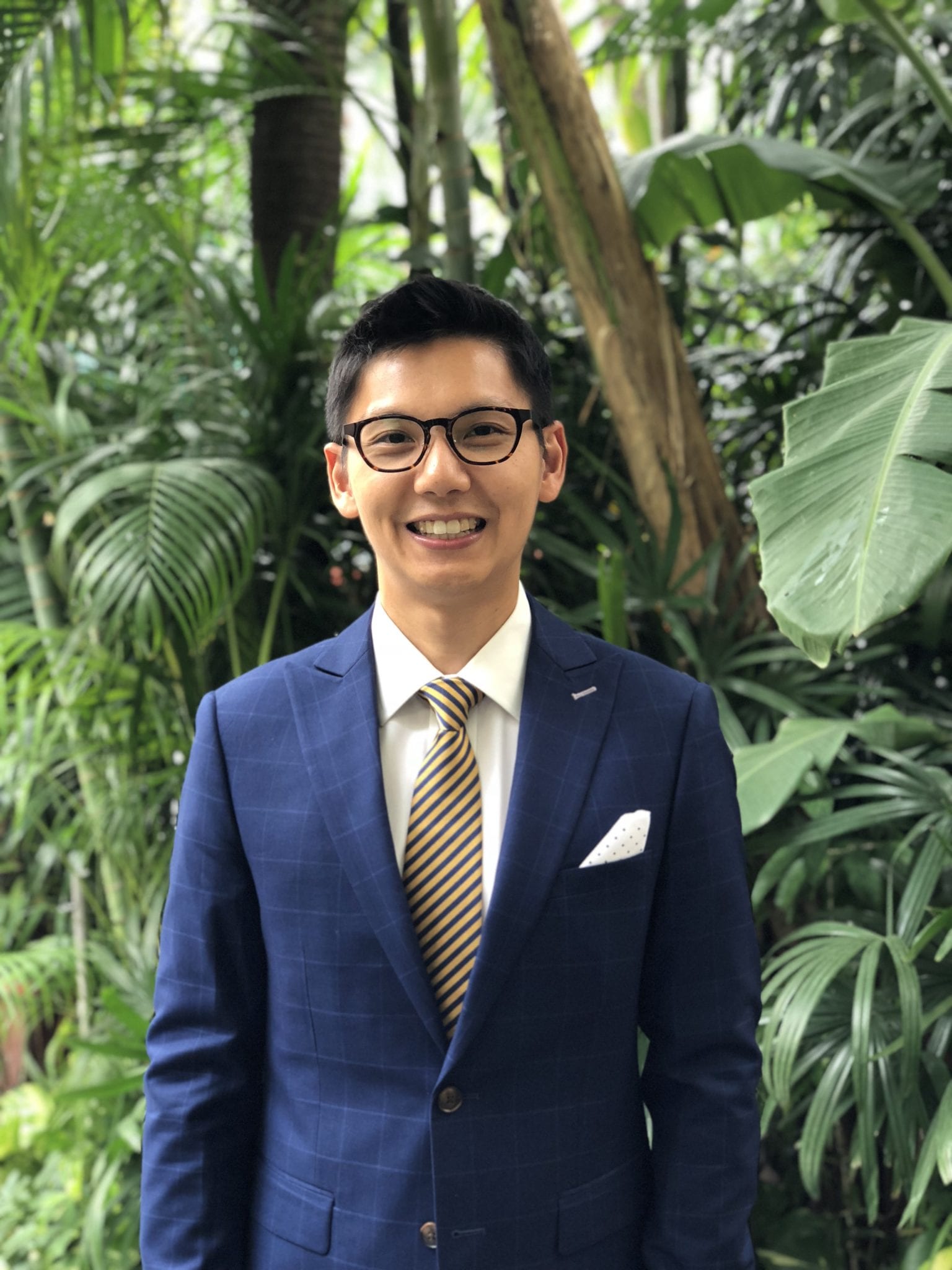 A headshot of smiling Chin-Hao Huang who has short black hair and black-rimmed glasses. He is wearing a tie with dark blue and yellow stripes, a white collared shirt under a navy-blue suit jacket, and has a white pocket square with polka dots.