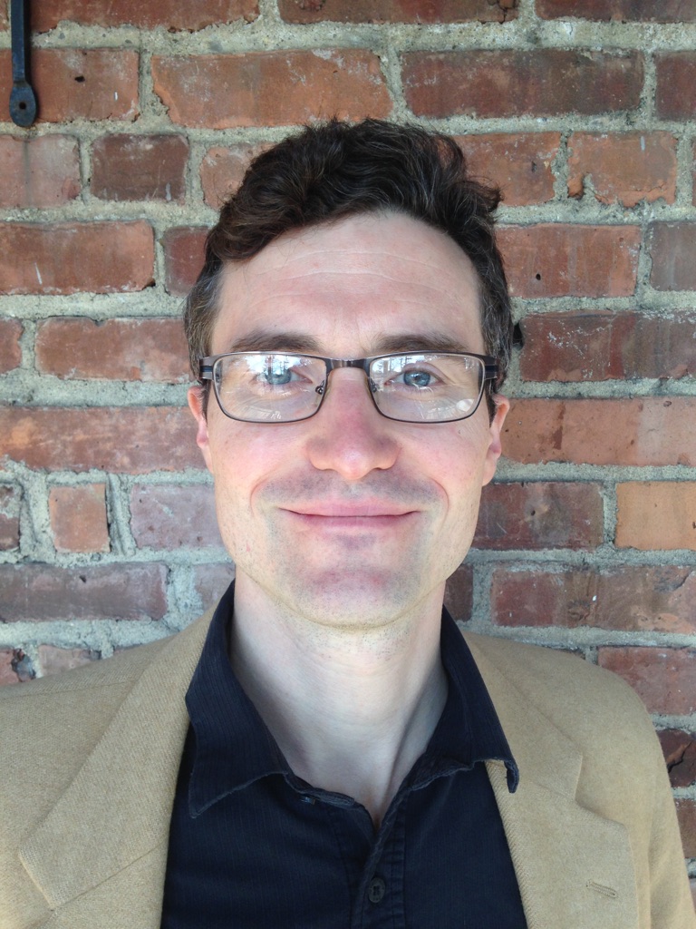 A headshot of smiling Stuart Earle Strange who has short brown hair, wearing glasses with black frame and a light brown blazer with a black collared shirt underneath. He is standing in front of a red brick wall.