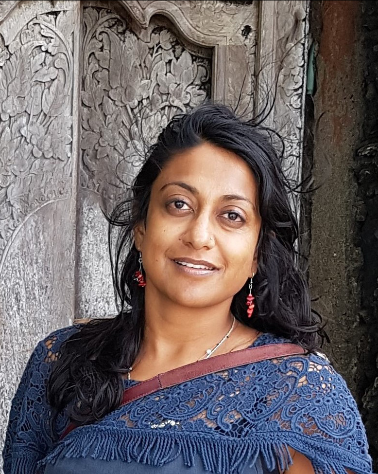 A headshot of smiling Neena Mahadev who has long black hair past her shoulders, wearing a sari made of blue lace with a maroon silk trim. She is standing in front of a wooden structure featuring intricate carvings.