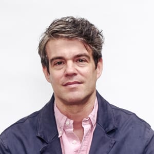 A headshot of Joshua Comaroff who has wavy brown hair. He is wearing a navy-blue jacket with a pink collared shirt underneath.