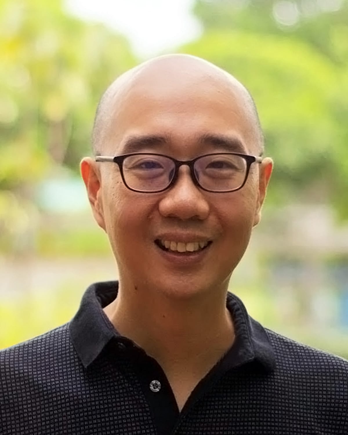 A headshot of smiling Marvin Joseph Fonacier Montefrio who is wearing glasses with a black frame and a black polo shirt with greenery in the background.