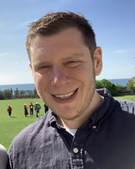 A headshot of smiling Justin D Stern who has short chestnut hair and is wearing a dark grey flannel shirt. He is standing on a green field; the ocean can be viewed in the background.