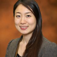 A headshot of smiling Suzuki Mao who has long brown hair past her shoulders, wearing a silver necklace and a grey blazer with a black blouse underneath. She is standing in front of a brown wall.