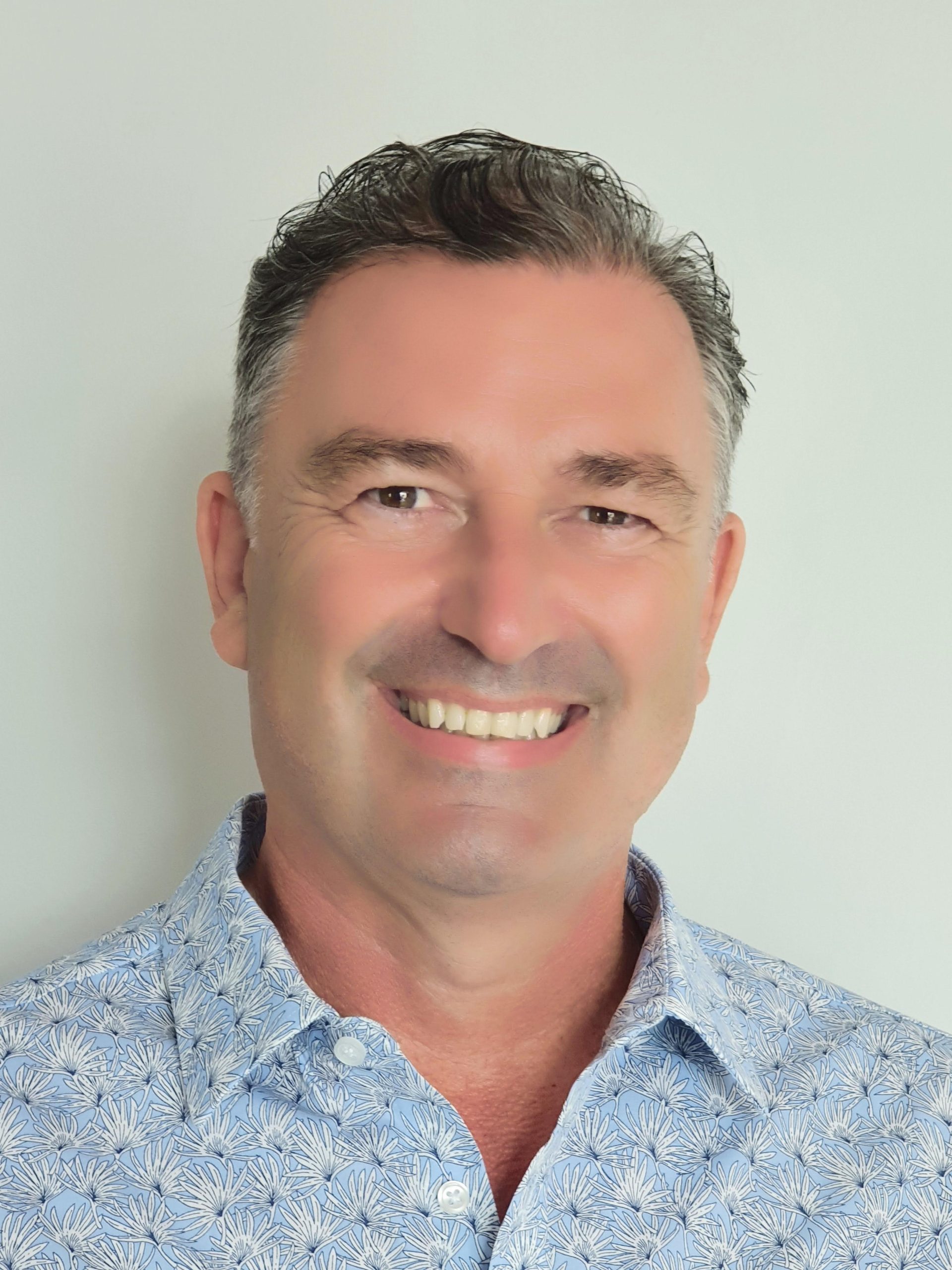 A headshot of smiling Stephen Brian Pointing who has short brown-grey hair. He is wearing a light blue collared shirt with a white flower pattern.