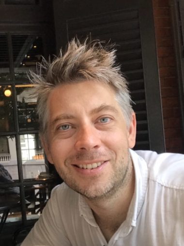 A headshot of smiling Benjamin Wainwright who has light-brown hair which is worn spiky and short. He is wearing a white collared shirt and is sitting in front of a window with grey shutters.