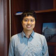 A headshot of smiling Michael Choi who has short black hair with a fringe, wearing a light blue collared shirt. He is standing in front of a large wooden bookshelf.