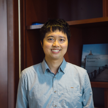 A headshot of smiling Michael Choi who has short black hair with a fringe, wearing a light blue collared shirt. He is standing in front of a large wooden bookshelf.
