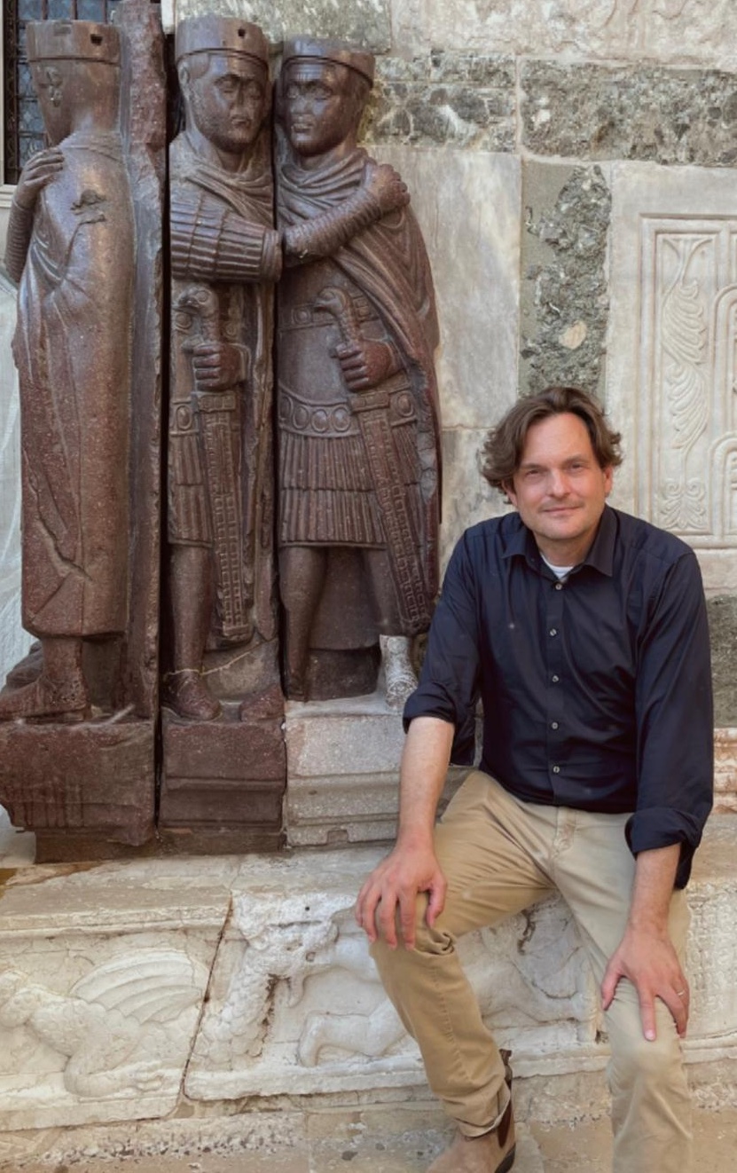 A headshot of smiling Ernst Emanuel Mayer who has short brown hair, wearing a blue shirt and brown pants. He is sitting in front of a statue.