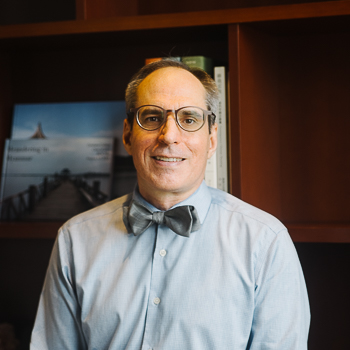 A headshot of smiling Paul Gallagher who has short brown-grey hair, wearing glasses with grey frames and a light blue collared shirt with a blue-grey bow tie on his neck. He is standing in front of wooden bookshelves.