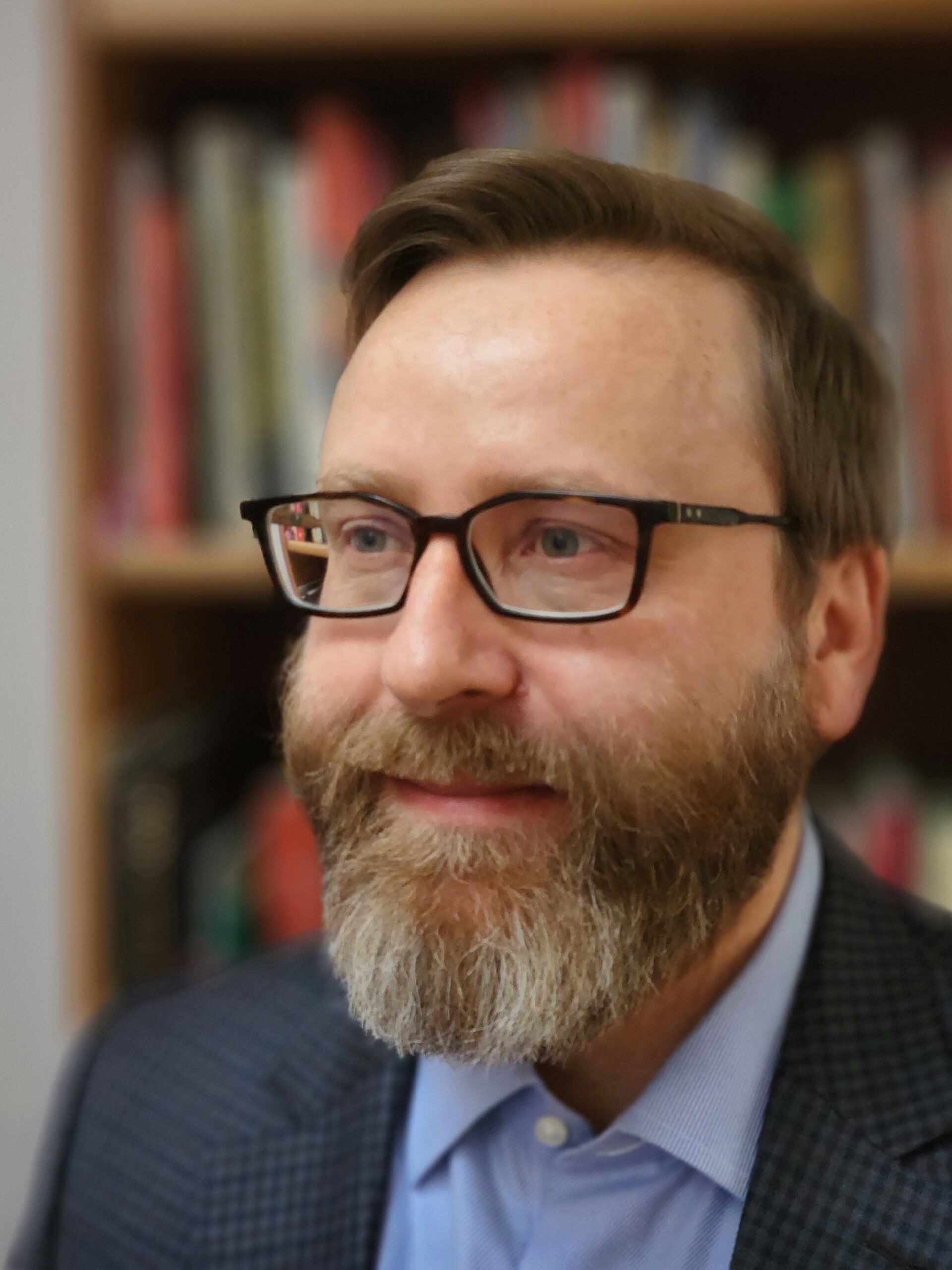 A headshot of smiling Zachary Moss Howlett who has brown hair, a mustache, and a beard. He is wearing glasses, a suit jacket with a dark grey and black checker pattern and a light blue collared shirt underneath.