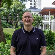 A headshot of smiling David Jacks who is wearing glasses and a dark blue polo shirt. There is greenery in the background.