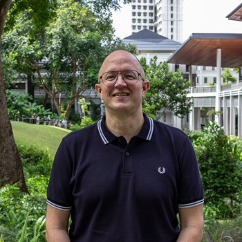 A headshot of smiling David Jacks who is wearing glasses and a dark blue polo shirt. There is greenery in the background.