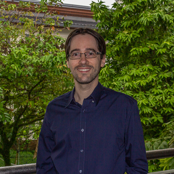 A headshot of smiling Sebastian Pohl who has short brown hair, a mustache, and a beard. He is wearing glasses with a black frame and a dark blue collared shirt with greenery in the background.