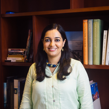 A headshot of smiling Smrithi Mohan Prasad who has long black hair past her shoulders, wearing leaf-shaped earrings and a large multi-stone necklace colored blue, green, and yellow. She is also wearing a light green long-sleeved blouse. She is standing in front of a large bookshelf.