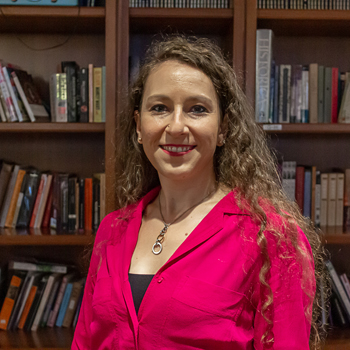 A headshot of smiling Francesca Spagnuolo who has wavy, long brown hair, and is wearing a magenta blouse and a necklace. She is standing in front of a large bookshelf.