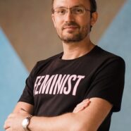 A headshot of smiling Stanislav Presolski who has short brown hair, a mustache, and a beard, wearing a black “Feminist” t-shirt. He is posing by crossing his hands over his chest, standing at the Yale-NUS Performance Hall foyer, in front of a wall drawing.