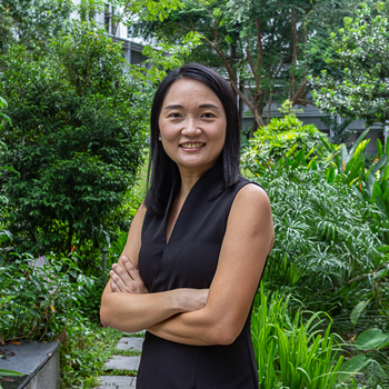 A headshot of smiling Eunice Jingmei Tan who has black hair of shoulder length, wearing a black V-neck dress. She is posing with her arms crossed in the Yale-NUS Campus Green.