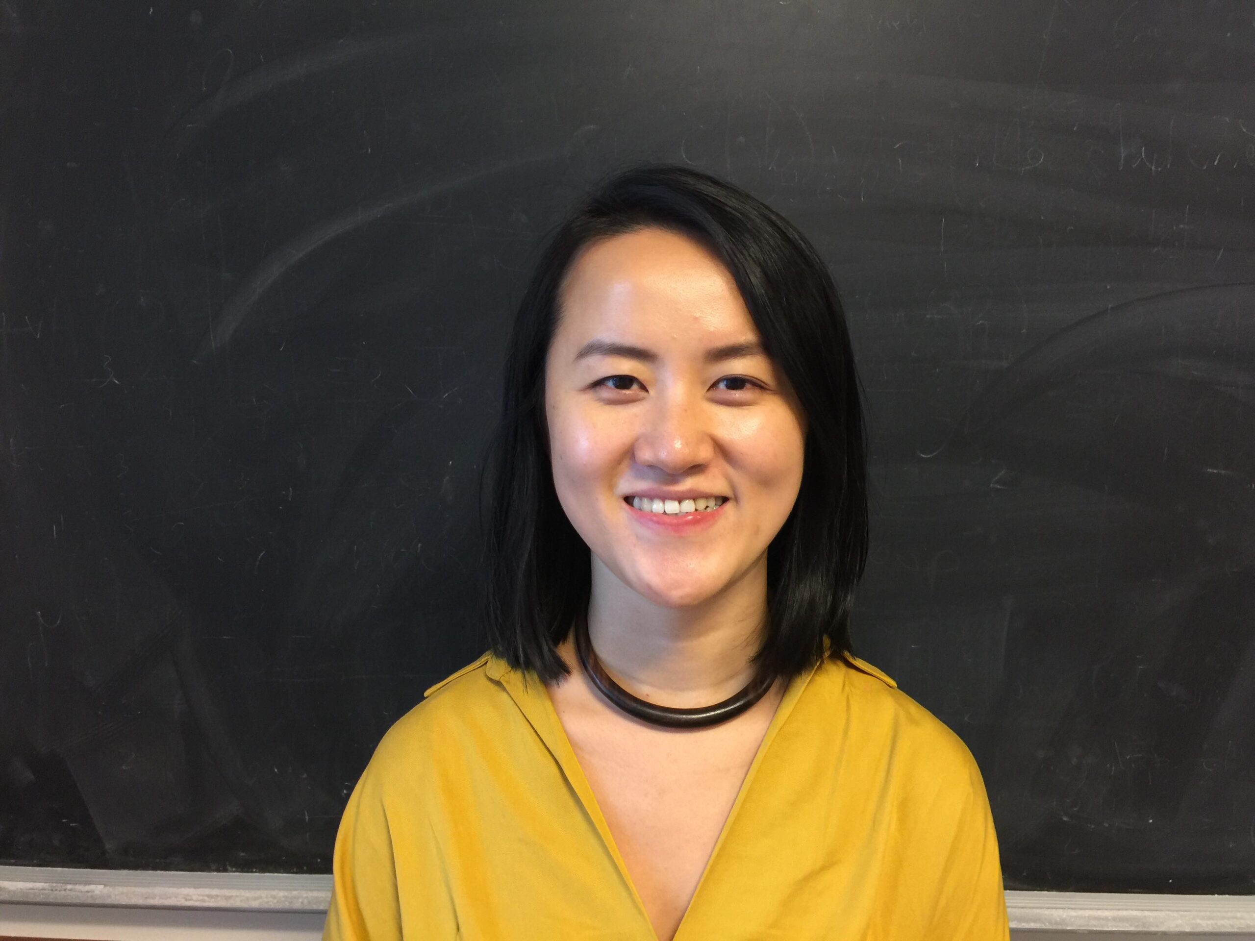 A headshot of smiling Xing Xia who has short black hair of shoulder length, wearing a yellow blouse and a black necklace with a black chalkboard in the background.