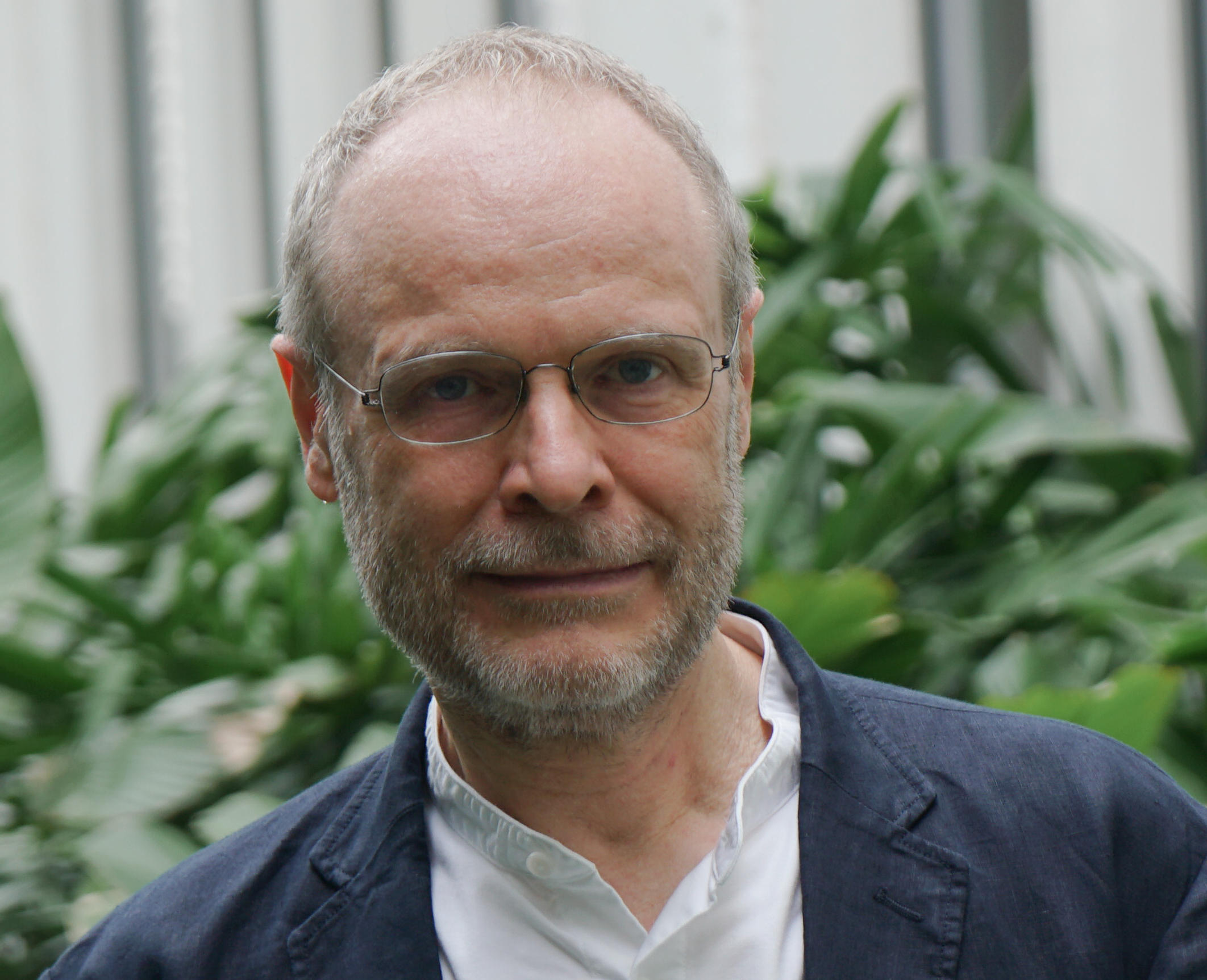 A headshot of smiling Olivier Danvy who has short white hair, a mustache, and a beard. He is wearing glasses with a grey frame and a blue collared jacket with a white collared shirt underneath. He is standing in front of greenery.