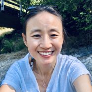 A headshot of smiling Yin Chang who has black hair in a ponytail. She is wearing sunglasses on top of her head, a gold necklace with a pearl, and a grey T-shirt. She is sitting on a rock with green plants and a bridge in the background.