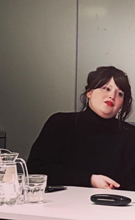 A headshot of smiling Swapna Kona Nayudu who has brown hair in a ponytail with middle-parted bangs. She is wearing bright red lipstick a black long sleeve turtleneck.