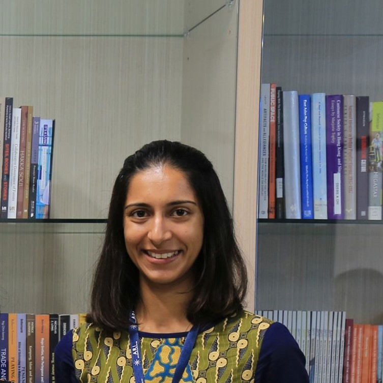A headshot of smiling Bittiandra Chand Somaiah who has short black hair of shoulder length, wearing an indigo long-sleeved collared shirt. She is standing in front of bookshelves.