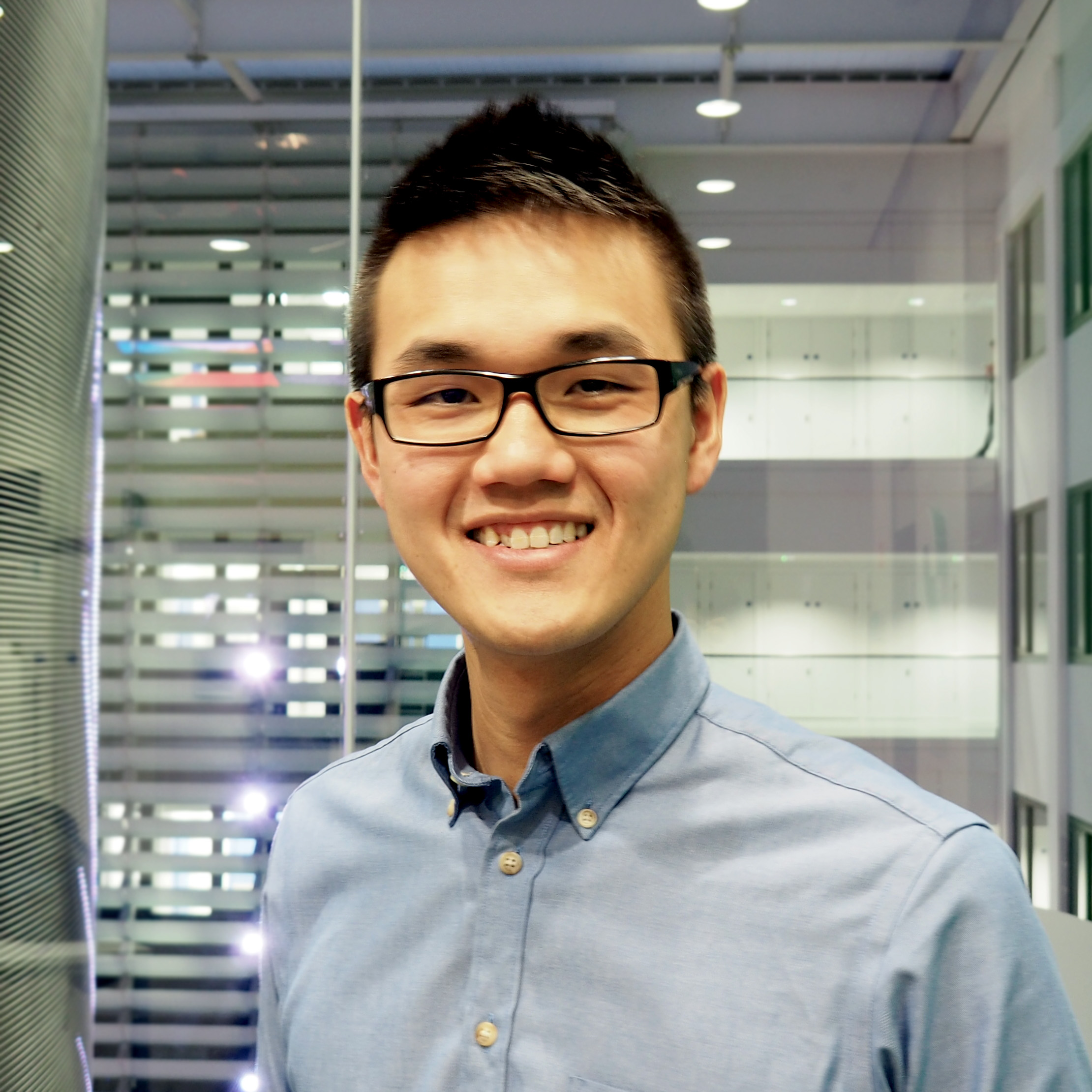 A headshot of smiling Zach Pang who has short black hair, wearing glasses with a black frame and a light blue collared shirt. He is standing in front of a glass window in an office, from which different levels of the of the building can be seen.